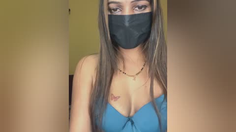 Video of a young woman with long brown hair, wearing a blue off-shoulder top and black face mask. She has a small butterfly tattoo on her chest and is standing indoors with a yellow wall in the background.
