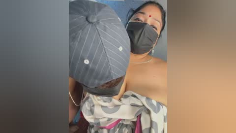 Video of a woman with medium brown skin, wearing a black mask, black headscarf, and patterned off-shoulder top, against a blurred blue and beige background.