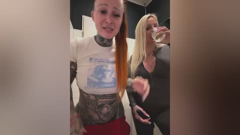 Media: Video of two women in a bathroom: one with a white \"Beverly Hills Cop\" t-shirt, heavily tattooed, and red pants; the other with blonde hair, black tank top, and drinking from a glass.