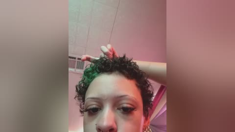 A close-up video of a young Black woman with curly hair, wearing a nose ring, and touching her hair with one hand. Background shows a tiled ceiling and pink wall.