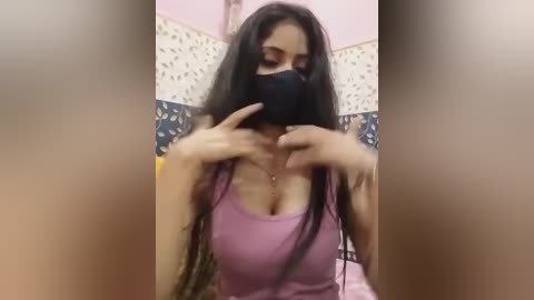 Media: A video of a young woman with long, dark hair, wearing a black mask and a low-cut pink tank top, adjusting her mask in a dimly lit room with floral wallpaper.