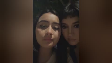 Video of two young women with dark hair, wearing black tops, lying close together, one with a pout, the other with a neutral expression, against a dark background.