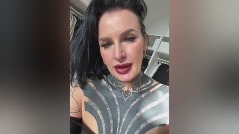 A close-up video of a pale-skinned woman with black hair, wearing dark lipstick, a silver body suit with intricate geometric patterns, and a nose ring. The background is blurry.