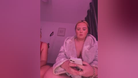 Video of a plus-sized woman with blonde hair, wearing a grey bathrobe, taking a selfie in a dimly lit bathroom with pinkish lighting, holding a smartphone.