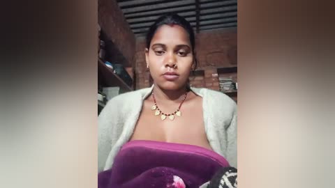 Video of a South Asian woman with medium brown skin, wearing a maroon sari with a gold necklace, sitting in a dimly lit room with a brick wall and wooden shelves.