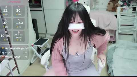 Media: Video of an Asian woman with long black hair, wearing a pink sweater and overalls, kneeling on a bed in a bedroom, with a virtual reality headset, displaying a video game interface.