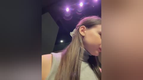 Media: A video of a woman with long, straight brown hair, partially visible. She is wearing a light grey turtleneck sweater and a white headband. The background features a dark, angular ceiling with purple lighting and a blurred figure in the background.