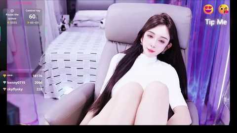 Video of an East Asian woman with long black hair, wearing a white turtleneck, sitting on a gray chair in a modern, dimly-lit bedroom.