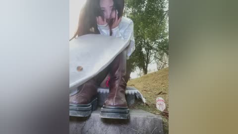Video of a woman with long brown hair crouching on a concrete ledge, wearing a white shirt and brown boots, holding a white board with a lens flare effect in the foreground.