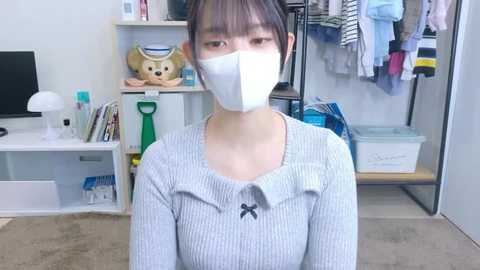 Video of an Asian woman with short dark hair, wearing a white mask, light gray ribbed sweater, and sitting on a beige carpet. Background includes shelves with stuffed animals, a white lamp, and hanging clothes.