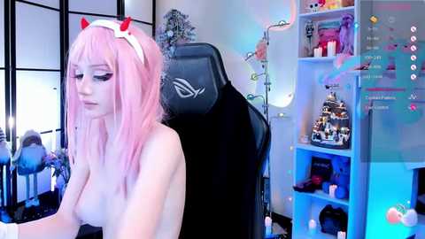 A video of a young, light-skinned woman with pink hair wearing cat ears, seated in a gaming chair, nude. Background features a desk with gaming accessories, a shelf with figurines, and a glowing screen displaying live chat.