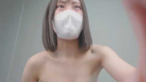 Video of an Asian woman with shoulder-length black hair, wearing a white surgical mask and no top, standing indoors with a plain background.