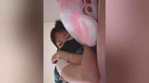 Video of an Asian woman in a pink room, wearing a blue dress, black mask, and white socks, lying on a bed, with a plush toy beside her.