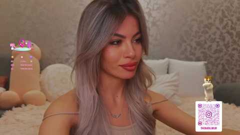 Video of a young woman with long, wavy, light brown hair, wearing a beige top, sitting on a bed with a plush teddy bear. Background features a patterned wallpaper and white pillows.