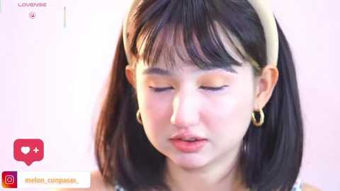 A video of a young Asian woman with straight black hair and a beige headband, wearing minimal makeup and gold hoop earrings, looking down with a serene expression.