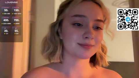 Video of a young Caucasian woman with shoulder-length blonde hair, smiling, wearing a strapless top. Background features a blurred indoor setting with a digital thermometer displaying 60\u00b0F, and a QR code in the top right corner.