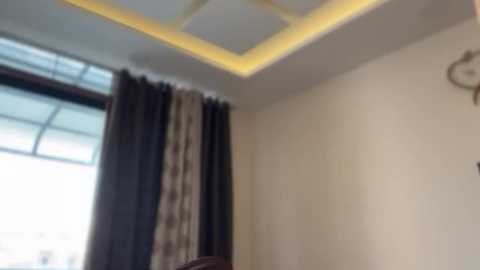 Video of a modern, brightly lit room with a recessed ceiling light, beige walls, dark curtains, and a window with a metal grid.