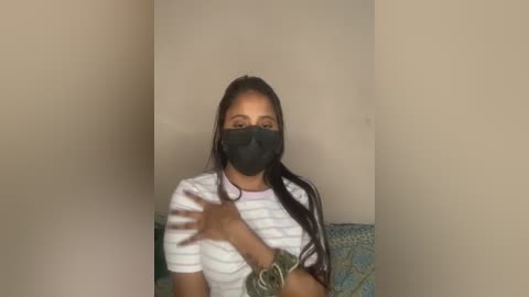 Video of a young woman with medium brown skin, wearing a black mask, white striped shirt, and a green bracelet, sitting against a beige wall, smiling.