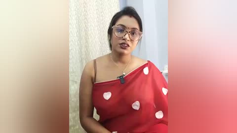 Video of a South Asian woman with medium brown skin, wearing a sheer red saree with white heart patterns, and large eyeglasses, standing against a textured wall.