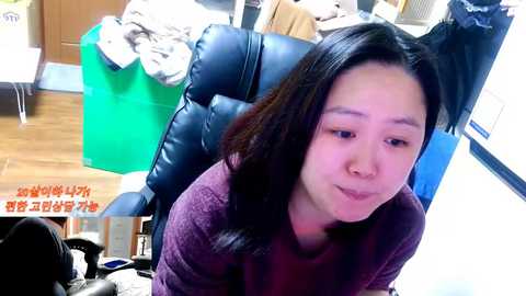 Video of an Asian woman with long black hair, wearing a maroon sweater, sitting in a black leather office chair in a cluttered room with a wooden floor.