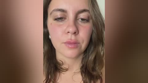 A close-up video of a young woman with fair skin, brown hair, and slightly puffy eyes, making a pouty face. The background is blurred, with a soft brown tone.