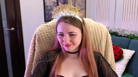 Video of a young, fair-skinned woman with long, straight light brown hair, wearing a golden crown, black choker, and a dark jacket. She smiles softly. Background includes a teal sofa, a beige blanket, and a red poinsettia.