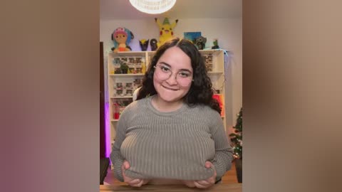 A video of a young woman with curly dark hair, glasses, and a gray ribbed sweater, holding her large breasts. Background features a Pikachu plush, colorful posters, and a bookshelf.