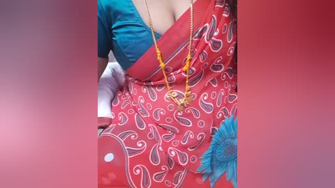 Video of a woman in a vibrant red saree with white paisley patterns and blue flowers, wearing a teal blouse, and a gold necklace with yellow beads, against a soft red background.
