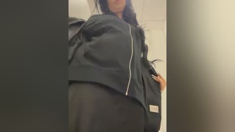 Video of a woman with long black hair, wearing a black jacket and black pants, standing in an indoor setting with neutral-colored walls and a ceiling.