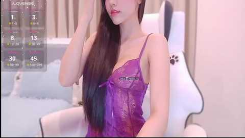Video of a young East Asian woman with long, straight black hair, wearing a sheer, purple lace lingerie bodysuit, seated on a white gaming chair in a softly lit, modern bedroom.