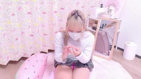 A video of a young Asian woman with blonde hair, wearing a white mask, white sweater, and short skirt, sitting on a pink, panda-patterned cushion, examining a smartphone. Background features a white wall, pink curtain, and a wooden shelf.