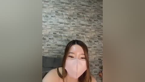 Video of a young Asian woman with long brown hair, wearing a pink face mask, standing in front of a grey stone wall, smiling.