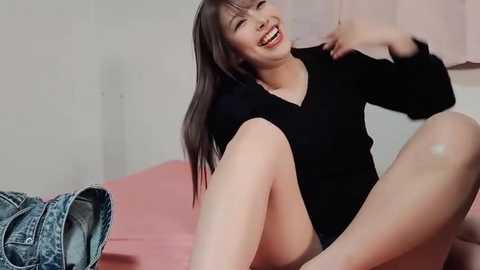 A joyful, smiling East Asian woman with long black hair sits on a pink bed, wearing a black top and shorts. She has a casual, denim jacket beside her.