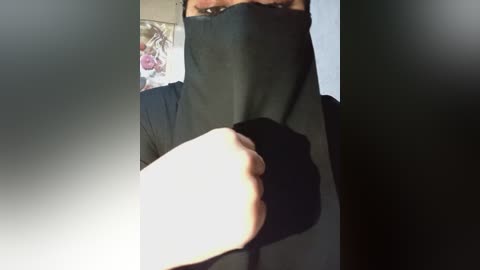 Video of a person wearing a black face mask and black hoodie, their hands clenched in front, in a dimly lit room with a wall-mounted painting in the background.