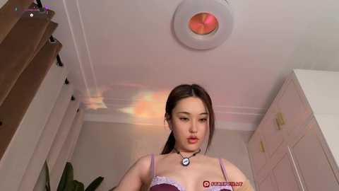 A video of an East Asian woman with long brown hair, wearing a purple bra and necklace, standing in a modern, minimalist room with white walls and a pink cabinet.