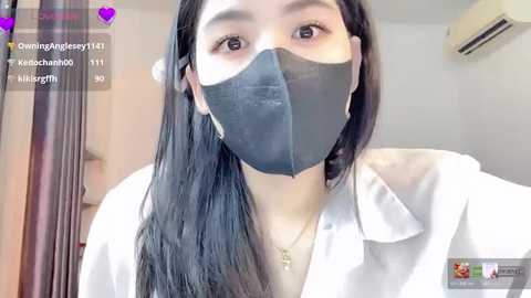 A video of an Asian woman with long black hair, wearing a black surgical mask and white shirt, taken indoors with a blurred background.