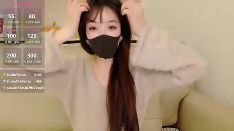 Media: A video of an East Asian woman with long black hair, wearing a black face mask and a beige cardigan, adjusting her hair in a living room with a beige couch and a TV displaying streaming stats.
