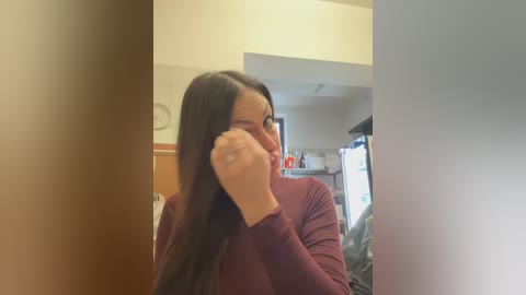 A video of a woman with long, straight brown hair, wearing a maroon long-sleeve top, covering her face with one hand, standing in a kitchen with beige walls, cabinets, and a refrigerator in the background.