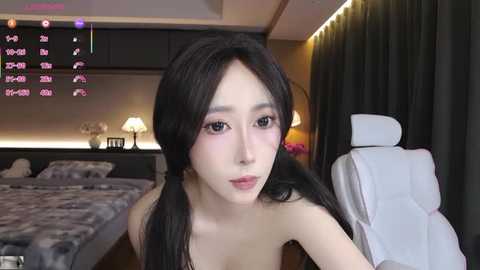 Video of a young Asian woman with long black hair, wearing a light pink lipstick, sitting on a white massage chair in a modern bedroom with a grey bed and pink floral decorations.