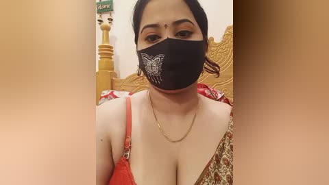 Media: Video of a South Asian woman with light skin, wearing a black face mask with a white design, a red dress, and a gold necklace, seated on a wooden bed with ornate carvings.