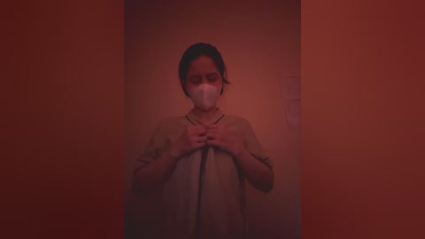 Video of a woman with dark hair, wearing a white surgical mask, standing in a dimly lit, narrow hallway with red-orange walls, hands clasped in front.