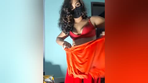 Video of a South Asian woman with dark curly hair, wearing a red bra and red pants, removing a red cloth, in a blue-walled room.