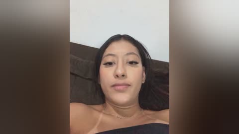 A video of a young Asian woman with long black hair, light skin, and a neutral expression, wearing a black off-shoulder top. She lies on a brown couch with a white wall behind her.