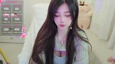 Video of an Asian woman with long black hair, fair skin, and a slender build, wearing a white, knitted sweater, seated on a bed. A stuffed pig is visible in the background.