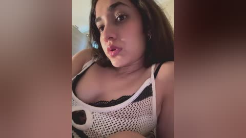 Video of a young woman with medium skin tone, dark hair, and full lips, wearing a white mesh top revealing black lace bra, indoors with blurred background.