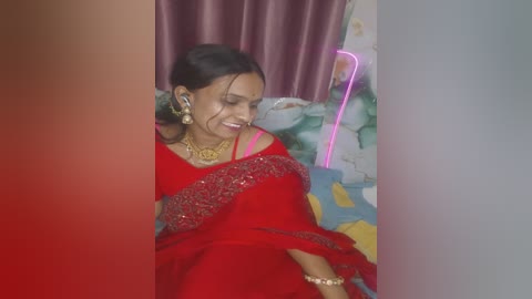Video of a South Asian woman in a red sari with gold embroidery, smiling, wearing gold jewelry, in a room with floral curtains and a colorful bedspread.
