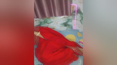 Video of a woman in a vibrant red sari with intricate gold embroidery, lying on a bed with a colorful, floral-patterned sheet. The background features a partially visible wall with a pinkish hue.