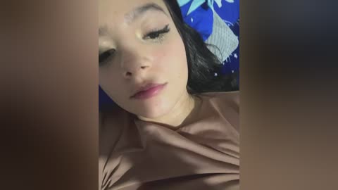 Video of a young Asian woman with long black hair, closed eyes, and subtle makeup, lying on a blue and white patterned pillow, wearing a pink shirt.