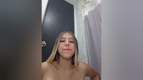 Video of a topless young woman with long blonde hair and fair skin, eyes closed, standing in a bathroom with a black door, white tiled walls, and a shower curtain.