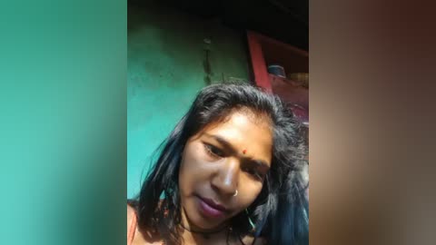 Video of a young woman with long black hair, wearing a pink top, against a teal and red wall.